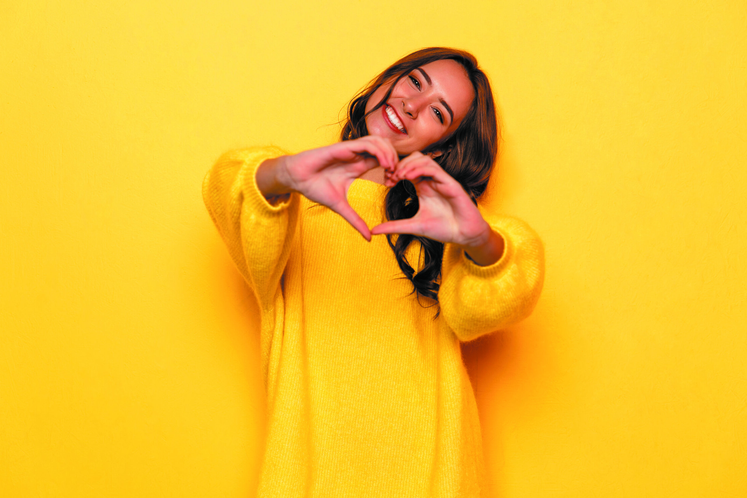 Smiling,young,girl,in,yellow,sweater,showing,heart,with,two